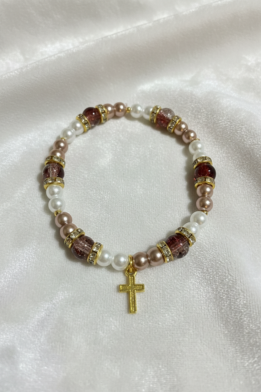 A beaded stretch bracelet featuring white faux pearls, dusty pink faux pearls, faceted reddish-brown beads, and gold spacer rings, with a small gold cross charm at the center, displayed on a white satin fabric background.