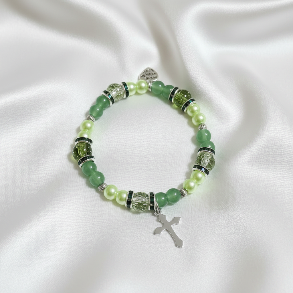 A beaded stretch bracelet featuring green and light green beads, clear faceted spacer beads, silver-toned metal spacers, a small silver heart charm engraved with 'JESUS IS LORD 230V', and a dangling gold-toned cross charm, shown on a plain white background.