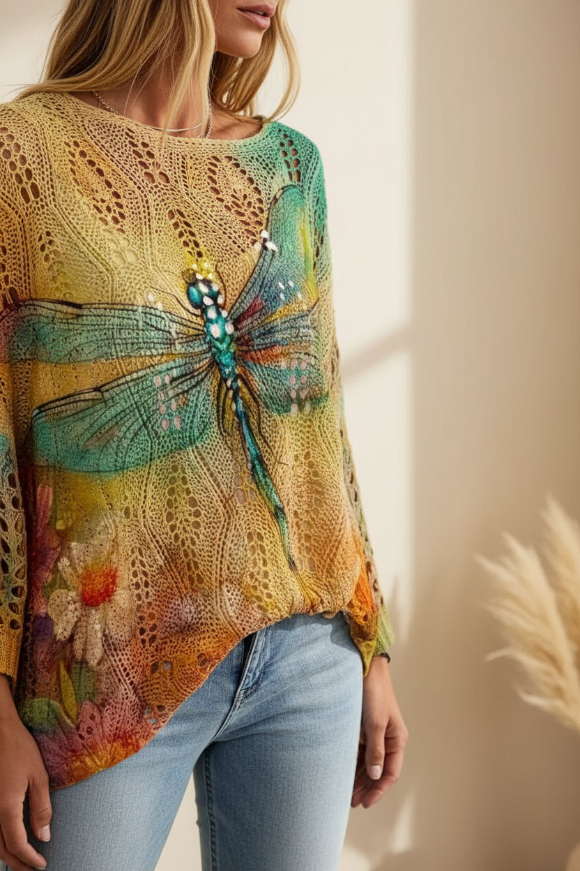 A close-up of a woman wearing a loose, short-sleeve crochet top with a large dragonfly design across the front, in earthy yellow, green, and orange tones, paired with blue jeans.