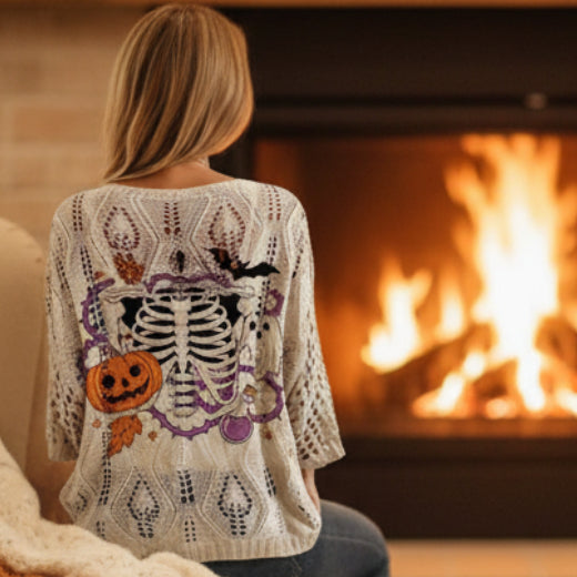 A woman wearing a cream-colored open-knit sweater with a large Halloween-themed back graphic featuring a skeleton, pumpkins, purple vines, and bats.