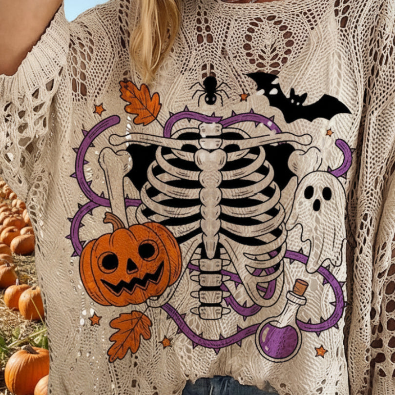 A woman wearing a cream-colored, open-knit long-sleeve sweater with a colorful Halloween graphic featuring a skeleton, pumpkin, ghost, bat, spider, and purple witch's hat.