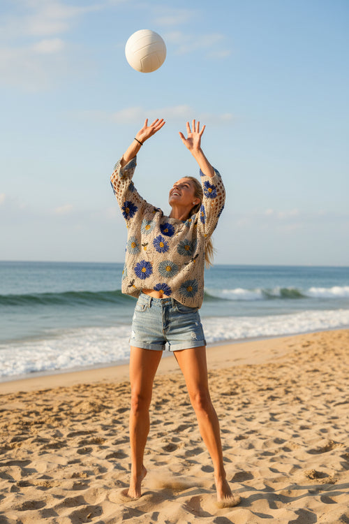 A woman wearing a beige crochet knit top with large blue and light blue floral motifs and small black-and-yellow butterfly details, paired with light blue jeans.
