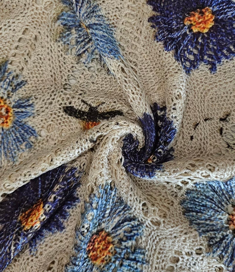 A close-up of a knitted fabric with a floral pattern, featuring blue and purple flowers with orange centers on a beige background.
