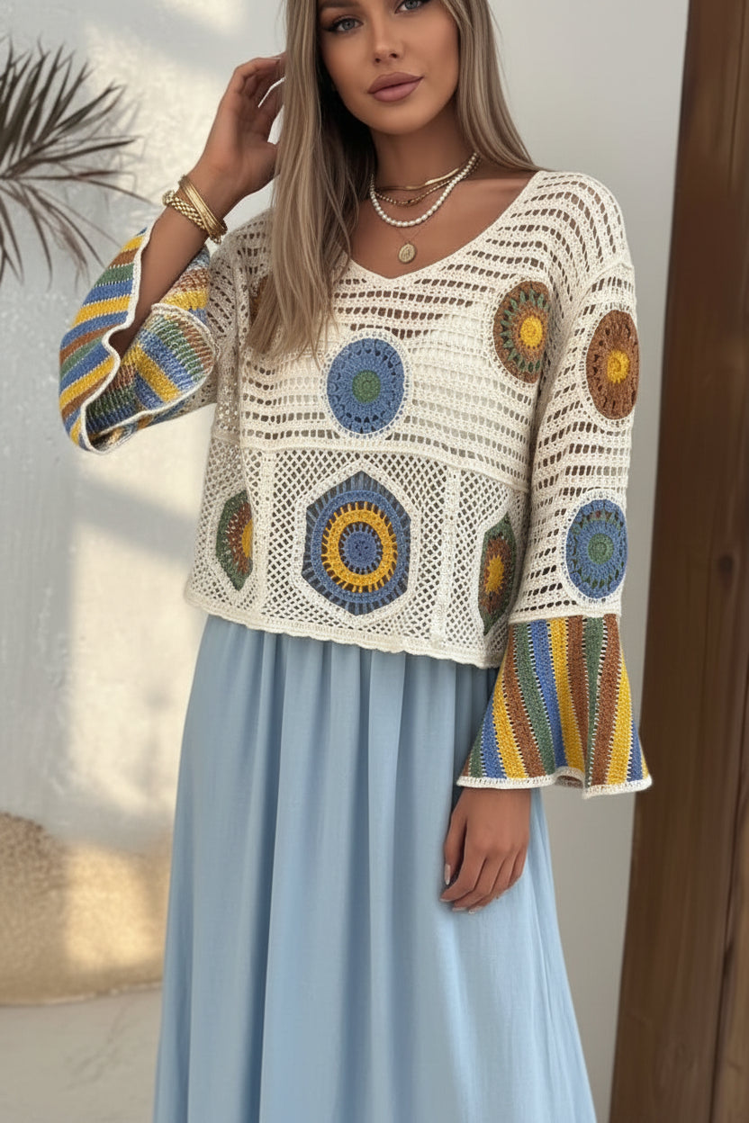 A woman wearing a cream crochet bohemian-style top with colorful circular granny square motifs and striped bell sleeves, paired with light-wash distressed denim shorts.