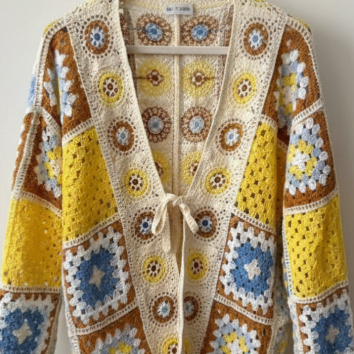 A handmade crocheted patchwork cardigan featuring yellow, blue, brown, and cream granny squares, held up by a hand on a metal chair.