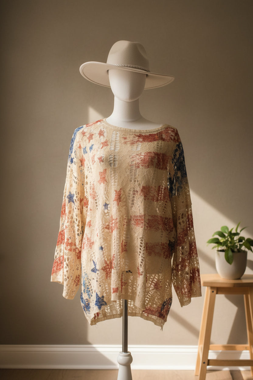 A long-sleeve crochet top with a colorful floral pattern on a cream base, displayed on a mannequin wearing a white wide-brim hat in a room with blue walls.