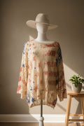 A long-sleeve crochet top with a colorful floral pattern on a cream base, displayed on a mannequin wearing a white wide-brim hat in a room with blue walls.