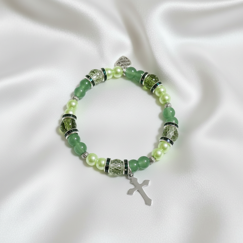 Green beaded stretch bracelet with pearl-style accents, silver-toned spacers, heart charm, and cross charm on white satin