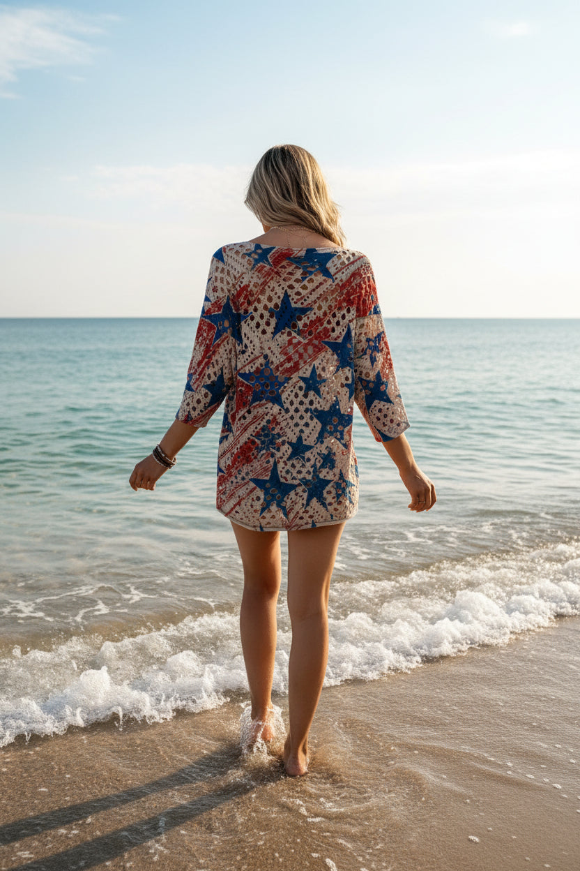 A woman shown from the back wearing a loose, open-knit top with an American flag-inspired pattern of red, white, and blue stars and stripes over a cream base, posed against a rustic white wooden wall.