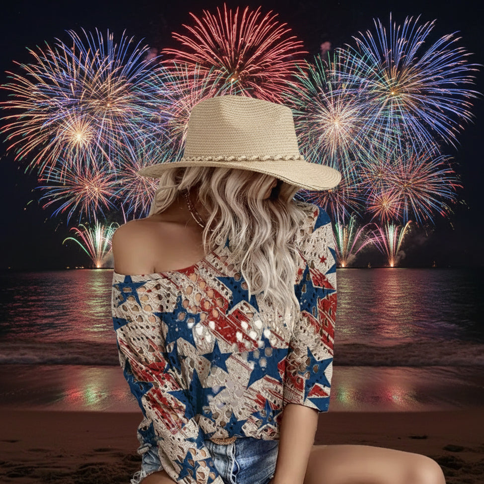 A woman wearing a patriotic off-the-shoulder top with a red, white, and blue American flag star pattern, paired with denim shorts and a straw hat, seated against a rustic wooden wall.